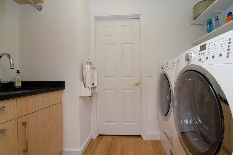Inredning av en modern liten svarta parallell svart tvättstuga enbart för tvätt, med en undermonterad diskho, släta luckor, skåp i ljust trä, bänkskiva i täljsten, vita väggar, mellanmörkt trägolv, en tvättmaskin och torktumlare bredvid varandra och orange golv