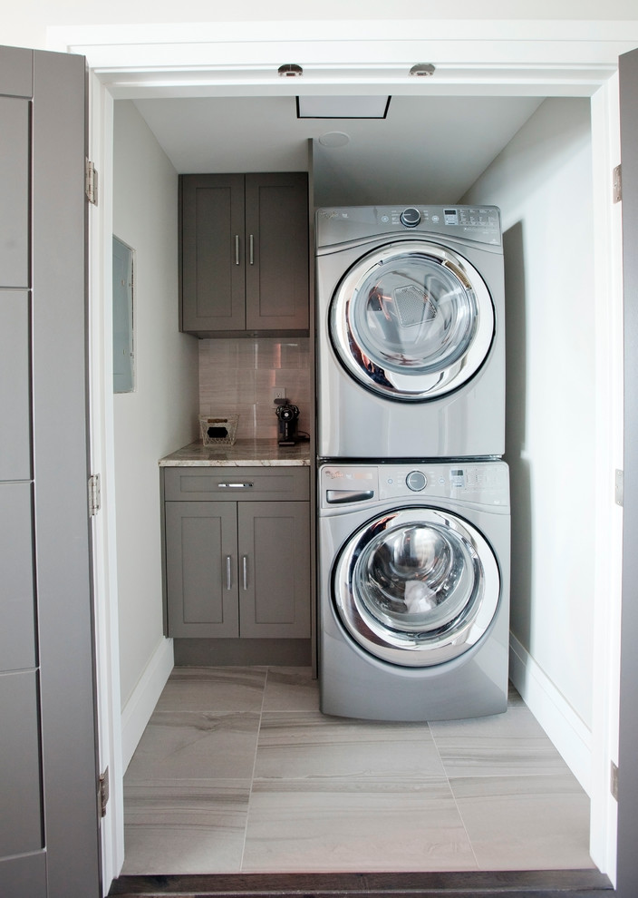Foto de lavadero lineal actual pequeño con armarios estilo shaker, puertas de armario grises, encimera de granito, paredes blancas, suelo de baldosas de porcelana y lavadora y secadora apiladas