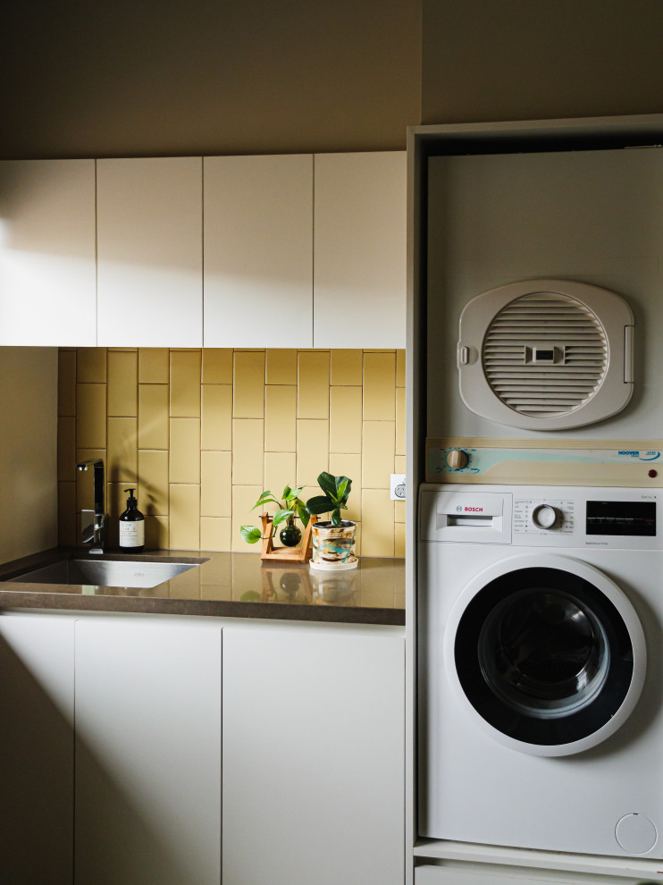 Foto på en mellanstor funkis bruna parallell tvättstuga enbart för tvätt, med släta luckor, stänkskydd i keramik, bänkskiva i kvarts, en undermonterad diskho, vita skåp, gult stänkskydd, skiffergolv, en tvättpelare och flerfärgat golv