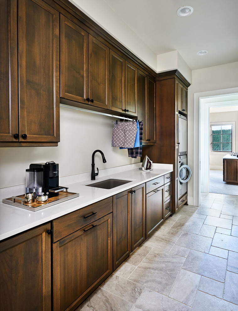 Inspiration for a large farmhouse single-wall limestone floor and beige floor laundry room remodel in Portland with an undermount sink, quartz countertops, white walls, a stacked washer/dryer, recessed-panel cabinets and dark wood cabinets