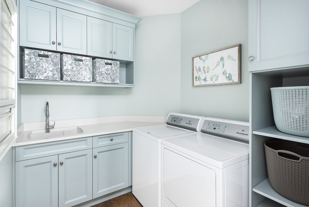 Bild på en stor vintage vita l-formad vitt tvättstuga enbart för tvätt, med skåp i shakerstil, blå skåp, bänkskiva i kvarts, blå väggar, en tvättmaskin och torktumlare bredvid varandra, brunt golv och mörkt trägolv