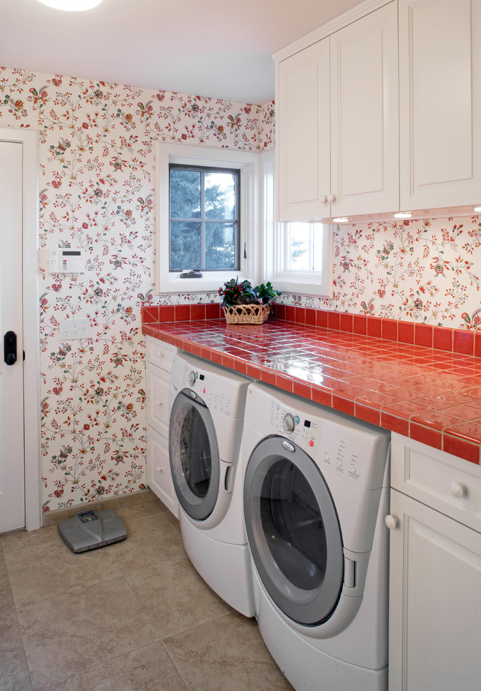 オマハにあるトラディショナルスタイルのおしゃれな洗濯室 (I型、レイズドパネル扉のキャビネット、白いキャビネット、タイルカウンター、マルチカラーの壁、左右配置の洗濯機・乾燥機、グレーの床、赤いキッチンカウンター) の写真