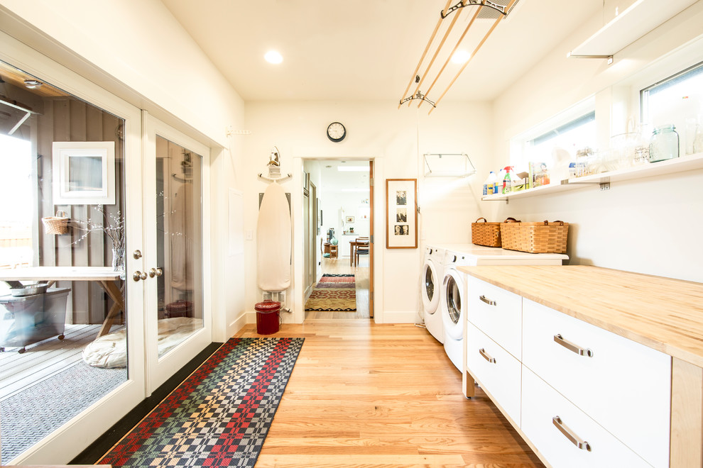 Einzeilige Moderne Waschküche mit flächenbündigen Schrankfronten, weißen Schränken, Arbeitsplatte aus Holz, weißer Wandfarbe, braunem Holzboden, Waschmaschine und Trockner nebeneinander, braunem Boden und beiger Arbeitsplatte in Seattle