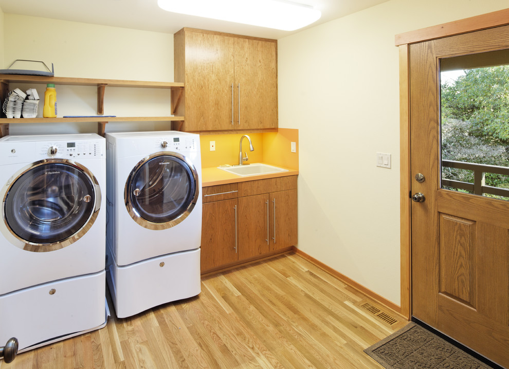 Idéer för att renovera en funkis tvättstuga enbart för tvätt, med en nedsänkt diskho, släta luckor, skåp i mellenmörkt trä, laminatbänkskiva, beige väggar, mellanmörkt trägolv och en tvättmaskin och torktumlare bredvid varandra