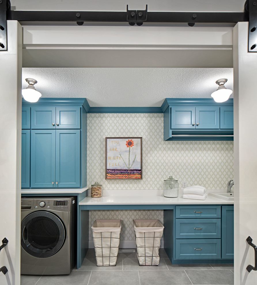 Inredning av en klassisk mellanstor linjär tvättstuga enbart för tvätt, med en nedsänkt diskho, blå skåp, bänkskiva i kvarts, klinkergolv i keramik, en tvättmaskin och torktumlare bredvid varandra, luckor med infälld panel och grå väggar