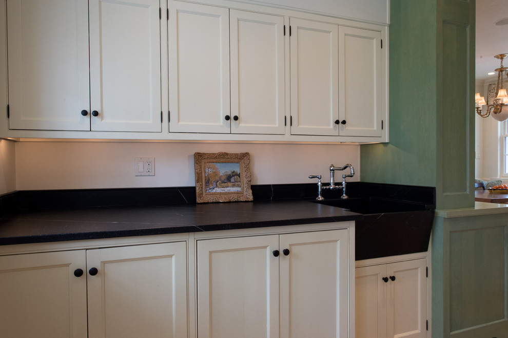 This is an example of a medium sized classic single-wall utility room in Philadelphia with a belfast sink, beaded cabinets, white cabinets, soapstone worktops, white walls, light hardwood flooring, a concealed washer and dryer, brown floors and black worktops.