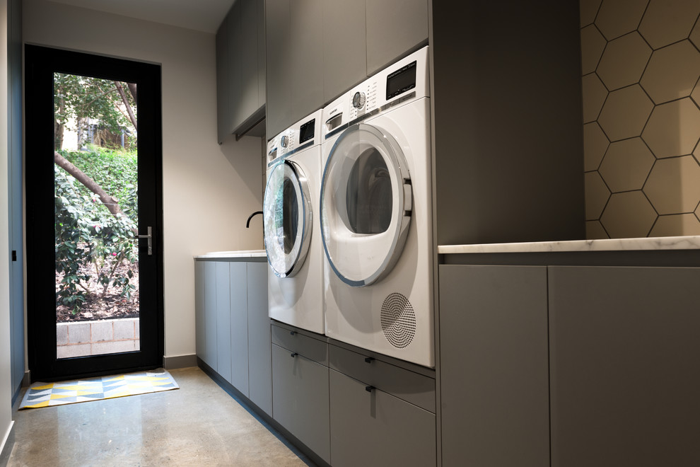 Foto de cuarto de lavado lineal contemporáneo de tamaño medio con fregadero encastrado, puertas de armario grises, encimera de cuarzo compacto, paredes blancas, suelo de cemento y lavadora y secadora juntas