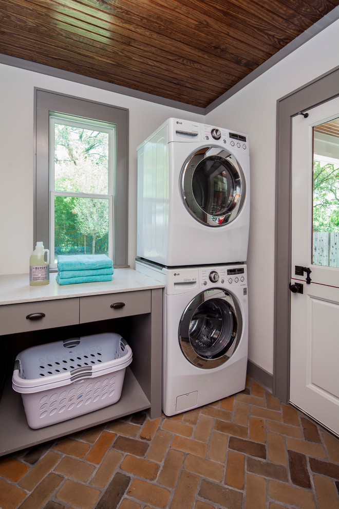 Photo of a traditional separated utility room in Austin with flat-panel cabinets, grey cabinets, engineered stone countertops, white walls, a stacked washer and dryer, brick flooring and white worktops.