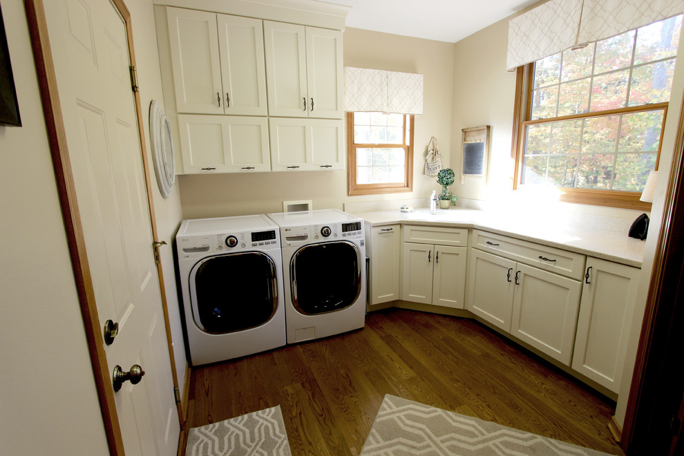 Mittelgroße Klassische Waschküche in L-Form mit Ausgussbecken, flächenbündigen Schrankfronten, dunklen Holzschränken, Mineralwerkstoff-Arbeitsplatte, beiger Wandfarbe, braunem Holzboden und Waschmaschine und Trockner nebeneinander in Cleveland