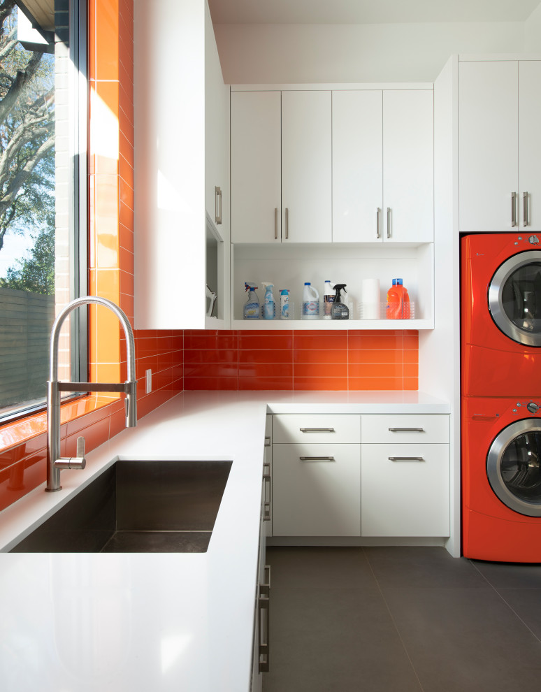 Modelo de cuarto de lavado en L contemporáneo grande con fregadero bajoencimera, armarios con paneles lisos, puertas de armario blancas, lavadora y secadora apiladas, suelo gris, encimeras blancas y salpicadero naranja