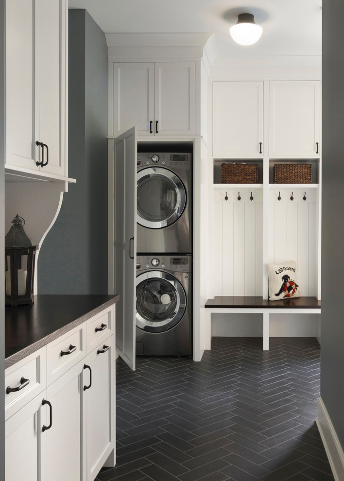 Modelo de lavadero multiusos costero con armarios estilo shaker, puertas de armario blancas, encimera de madera, paredes grises, lavadora y secadora apiladas y suelo negro