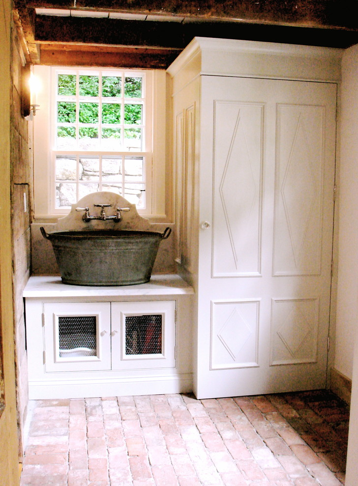 Exempel på en liten lantlig linjär tvättstuga, med en allbänk, luckor med profilerade fronter, vita skåp, marmorbänkskiva, vita väggar, tegelgolv och tvättmaskin och torktumlare byggt in i ett skåp