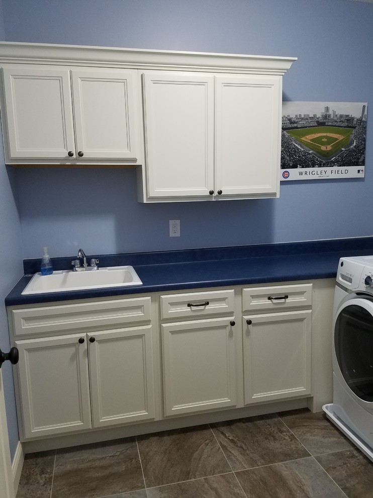 シカゴにある中くらいなおしゃれな洗濯室 (L型、ドロップインシンク、落し込みパネル扉のキャビネット、白いキャビネット、ラミネートカウンター、青い壁、セラミックタイルの床、左右配置の洗濯機・乾燥機、茶色い床、青いキッチンカウンター) の写真