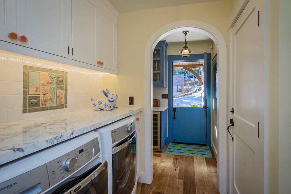 Einzeiliger Landhaus Hauswirtschaftsraum mit Schrankfronten im Shaker-Stil, weißen Schränken, Marmor-Arbeitsplatte, braunem Holzboden, Waschmaschine und Trockner nebeneinander und gelber Wandfarbe in San Francisco