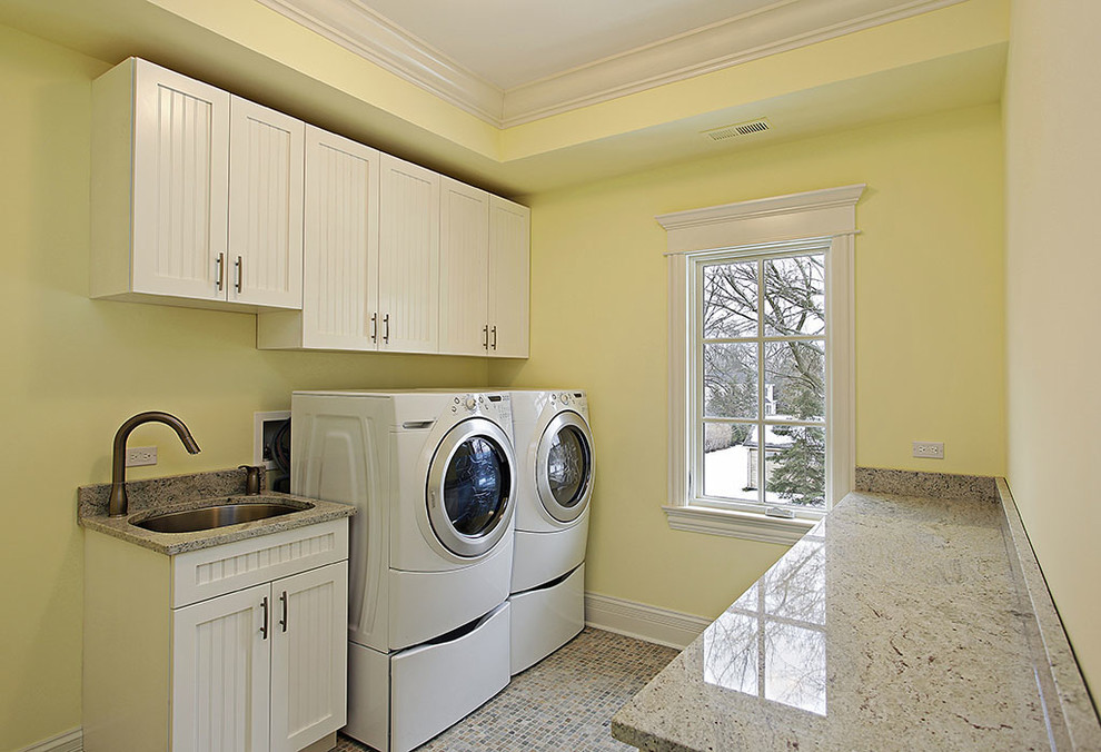 Exempel på en mellanstor klassisk bruna parallell brunt tvättstuga enbart för tvätt, med en undermonterad diskho, luckor med infälld panel, vita skåp, granitbänkskiva, gula väggar, en tvättmaskin och torktumlare bredvid varandra och flerfärgat golv