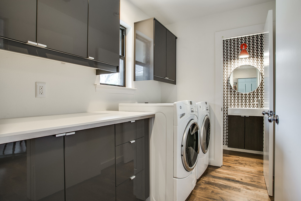 Bild på en funkis parallell tvättstuga, med släta luckor, bänkskiva i kvarts, vita väggar, mellanmörkt trägolv, en tvättmaskin och torktumlare bredvid varandra och grå skåp