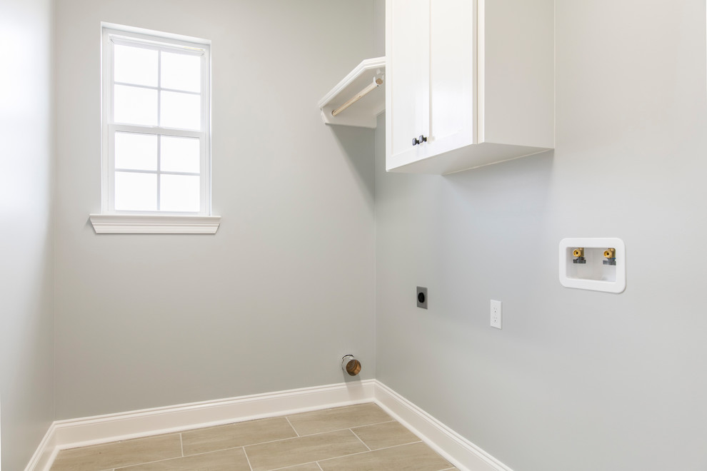 Modelo de cuarto de lavado lineal tradicional con armarios con paneles lisos, puertas de armario blancas, paredes grises, suelo de baldosas de cerámica, lavadora y secadora juntas y suelo gris