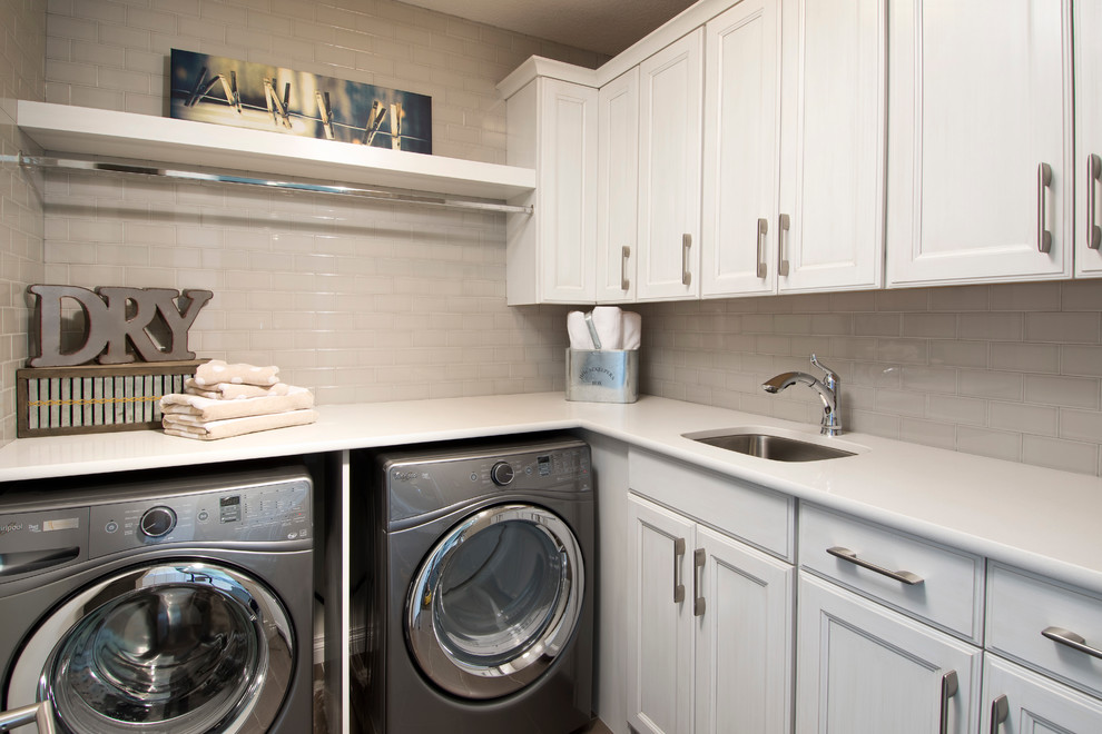 Inredning av en modern mellanstor l-formad tvättstuga enbart för tvätt, med en undermonterad diskho, luckor med profilerade fronter, vita skåp, laminatbänkskiva, vita väggar och en tvättmaskin och torktumlare bredvid varandra