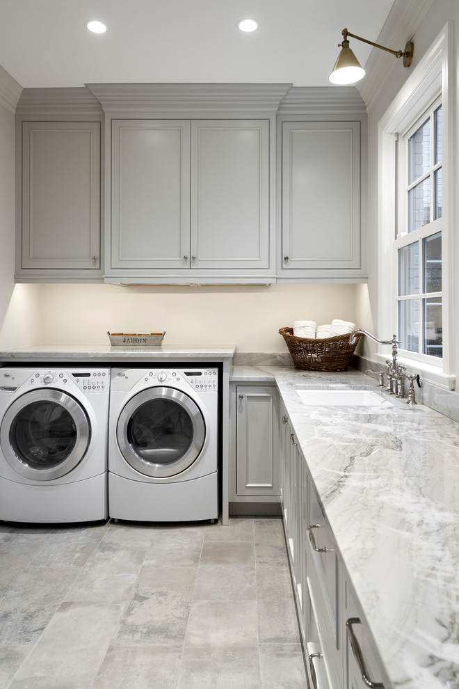 Inredning av en retro stor grå l-formad grått tvättstuga enbart för tvätt, med en undermonterad diskho, skåp i shakerstil, grå skåp, granitbänkskiva, beige väggar, klinkergolv i porslin, en tvättmaskin och torktumlare bredvid varandra och grått golv