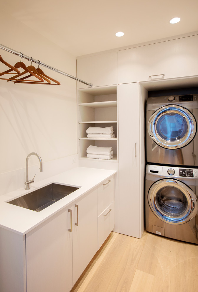 Small contemporary l-shaped separated utility room in Vancouver with a submerged sink, flat-panel cabinets, white cabinets, engineered stone countertops, white walls, light hardwood flooring, a stacked washer and dryer, white worktops and beige floors.