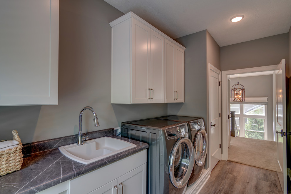 Inspiration för amerikanska linjära grått tvättstugor enbart för tvätt, med en nedsänkt diskho, vita skåp, laminatbänkskiva, grå väggar, vinylgolv, en tvättmaskin och torktumlare bredvid varandra och brunt golv