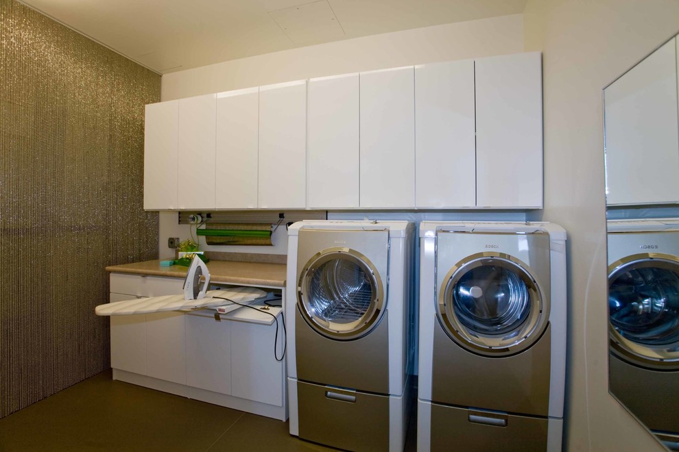 Modern inredning av en tvättstuga, med släta luckor, vita skåp, laminatbänkskiva, vita väggar, skiffergolv och en tvättmaskin och torktumlare bredvid varandra