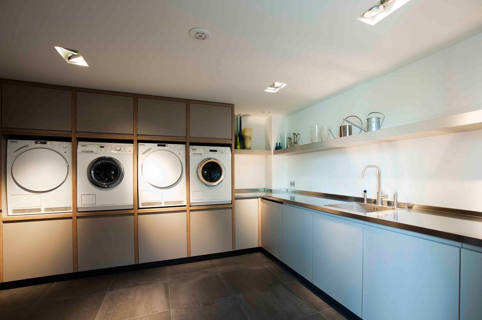 Modelo de lavadero multiusos y en L contemporáneo grande con fregadero integrado, encimera de acero inoxidable, paredes blancas, lavadora y secadora juntas, armarios con paneles lisos y puertas de armario azules