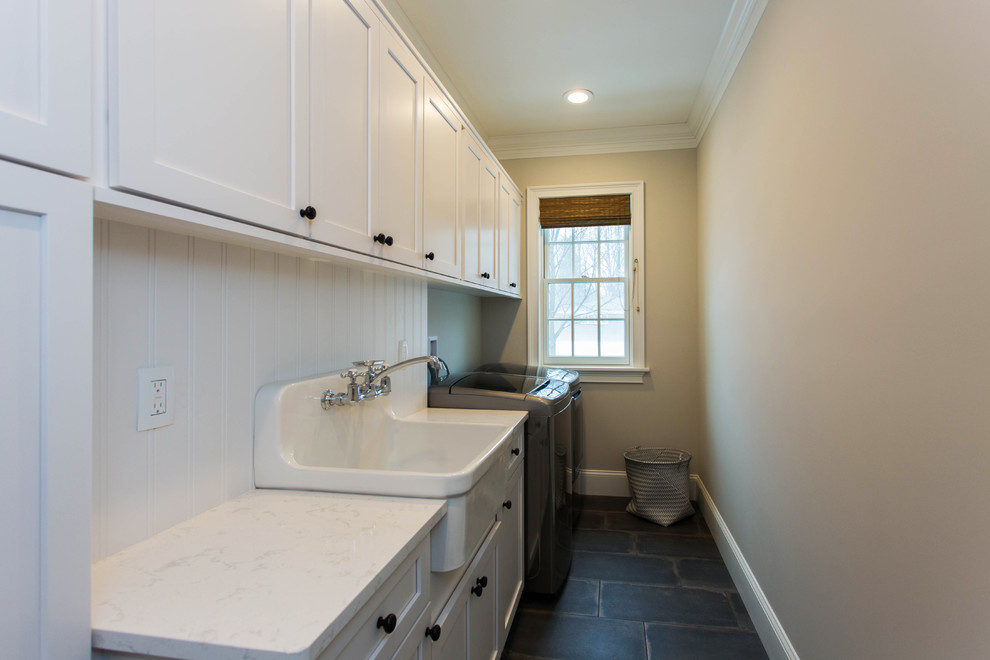 Klassisk inredning av en mellanstor linjär tvättstuga enbart för tvätt, med luckor med infälld panel, vita skåp, bänkskiva i kvarts, en tvättmaskin och torktumlare bredvid varandra, en rustik diskho, beige väggar, betonggolv och grått golv