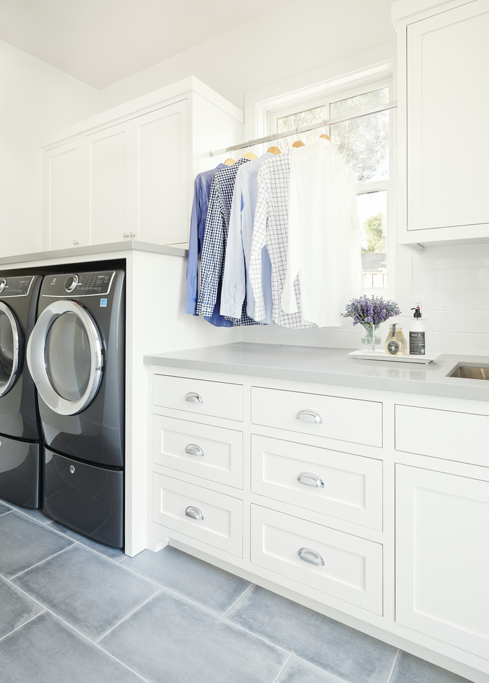 Lantlig inredning av en grå linjär grått tvättstuga enbart för tvätt, med en undermonterad diskho, skåp i shakerstil, vita skåp, bänkskiva i kvarts, vita väggar, klinkergolv i porslin, en tvättmaskin och torktumlare bredvid varandra och grått golv