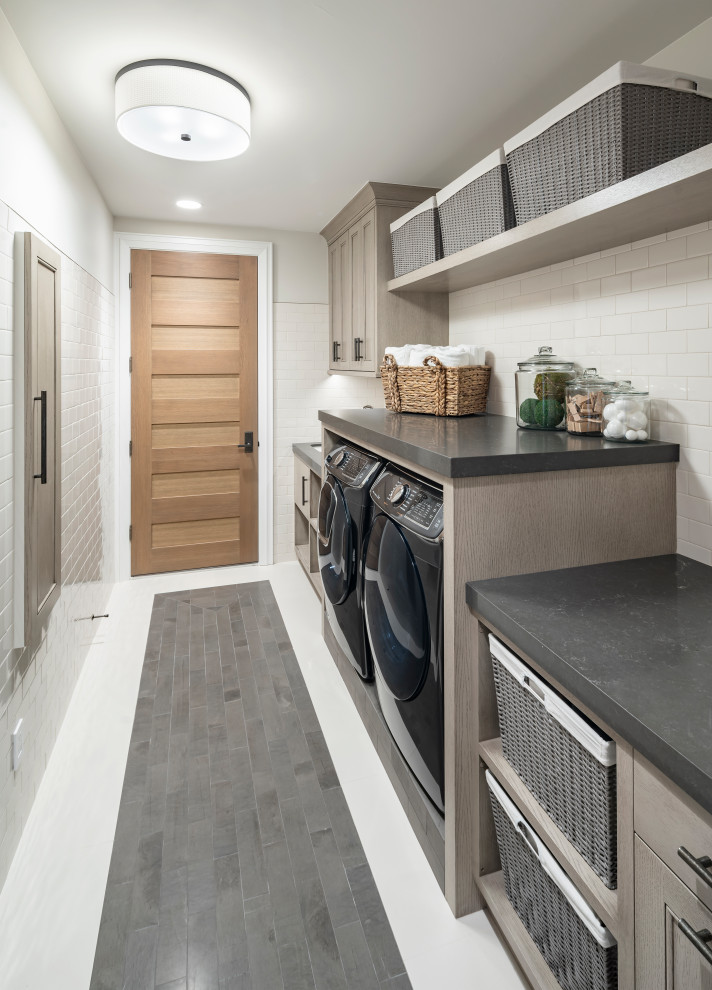 Bild på en stor lantlig grå parallell grått tvättstuga enbart för tvätt, med granitbänkskiva, skiffergolv, en tvättmaskin och torktumlare bredvid varandra, grått golv, luckor med infälld panel, grå skåp och grå väggar