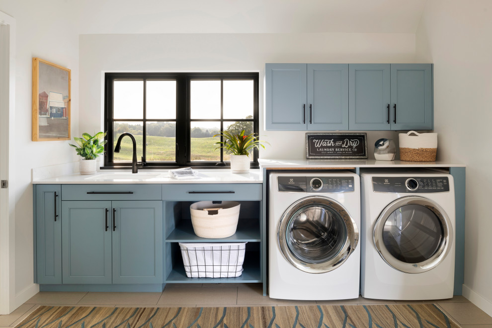 Imagen de lavadero multiusos y lineal costero grande con fregadero encastrado, puertas de armario azules, paredes blancas, suelo de madera clara, lavadora y secadora juntas y encimeras blancas
