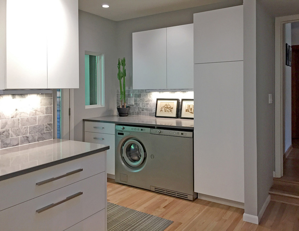 Photo of a small contemporary single-wall utility room in Milwaukee with flat-panel cabinets, white cabinets, engineered stone countertops, grey walls, light hardwood flooring and a side by side washer and dryer.
