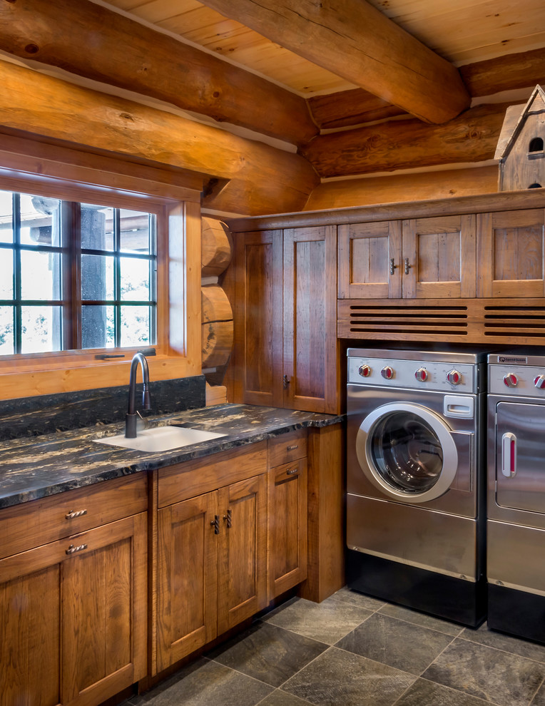 Bild på en stor rustik l-formad tvättstuga enbart för tvätt, med en undermonterad diskho, luckor med infälld panel, skåp i mellenmörkt trä, granitbänkskiva, skiffergolv och en tvättmaskin och torktumlare bredvid varandra