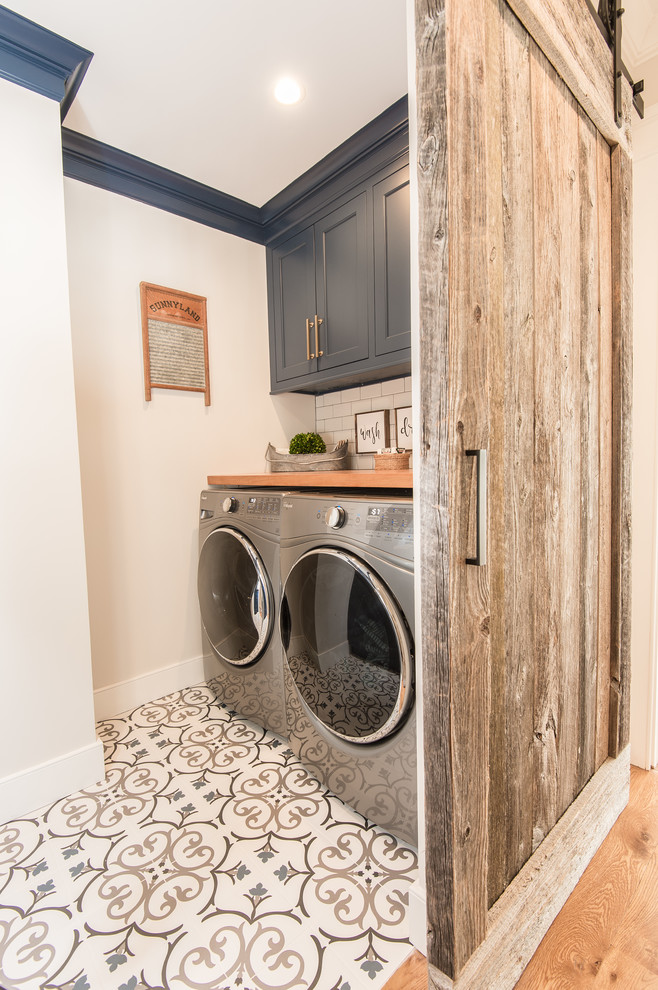 ブリッジポートにある高級な中くらいなカントリー風のおしゃれな洗濯室 (ll型、インセット扉のキャビネット、青いキャビネット、木材カウンター、白い壁、セラミックタイルの床、左右配置の洗濯機・乾燥機、白い床、茶色いキッチンカウンター) の写真