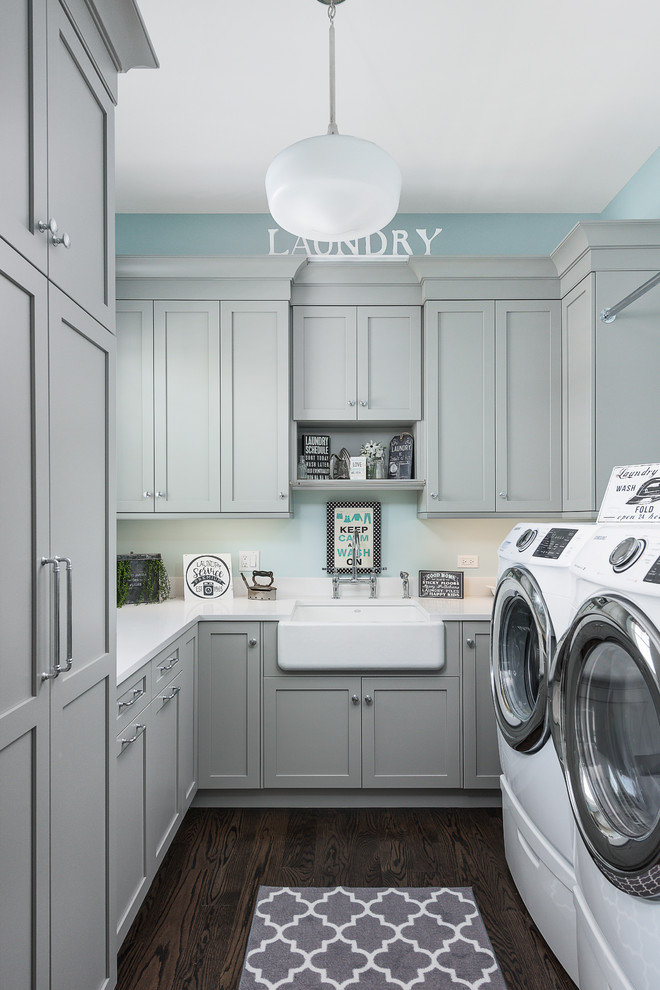 Klassische Waschküche in L-Form mit Landhausspüle, Schrankfronten im Shaker-Stil, grauen Schränken, blauer Wandfarbe, dunklem Holzboden, Waschmaschine und Trockner nebeneinander, braunem Boden und weißer Arbeitsplatte in Chicago