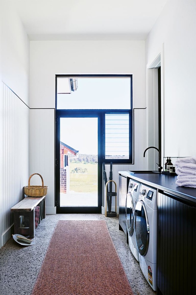 Exempel på ett stort lantligt linjärt grovkök, med en nedsänkt diskho, svarta skåp, laminatbänkskiva, vita väggar, betonggolv, en tvättmaskin och torktumlare bredvid varandra, grått golv och släta luckor