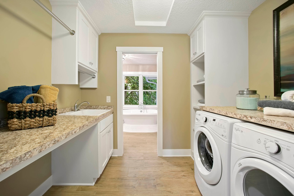 Große Landhaus Waschküche mit Landhausspüle, weißen Schränken, Granit-Arbeitsplatte, beiger Wandfarbe, braunem Holzboden und Waschmaschine und Trockner nebeneinander in Minneapolis