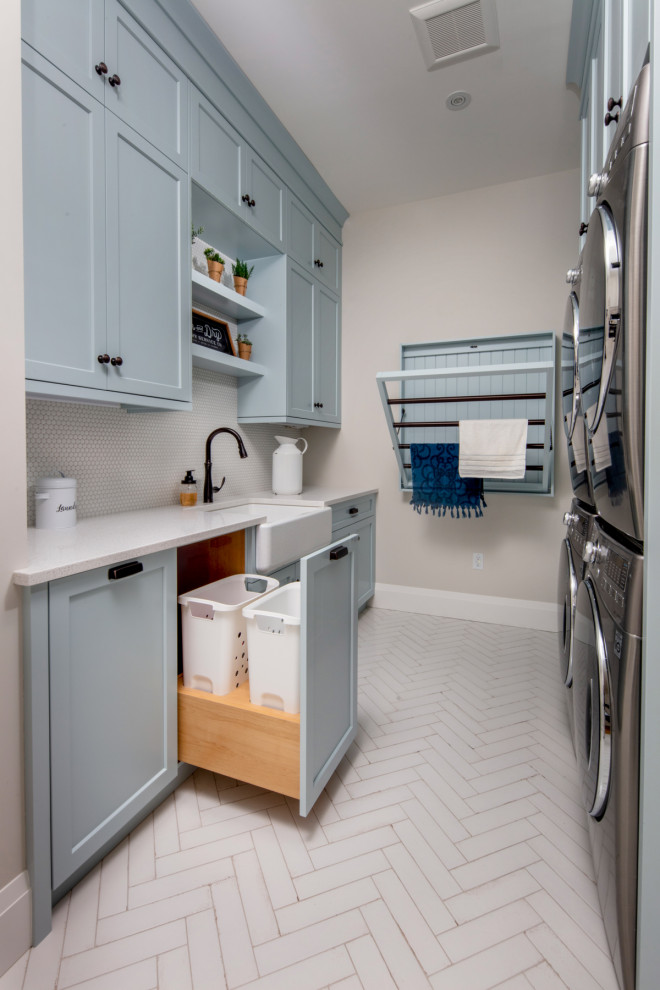 トロントにある中くらいなカントリー風のおしゃれな洗濯室 (ll型、エプロンフロントシンク、シェーカースタイル扉のキャビネット、青いキャビネット、クオーツストーンカウンター、グレーのキッチンパネル、セラミックタイルのキッチンパネル、ベージュの壁、上下配置の洗濯機・乾燥機、白い床、白いキッチンカウンター) の写真