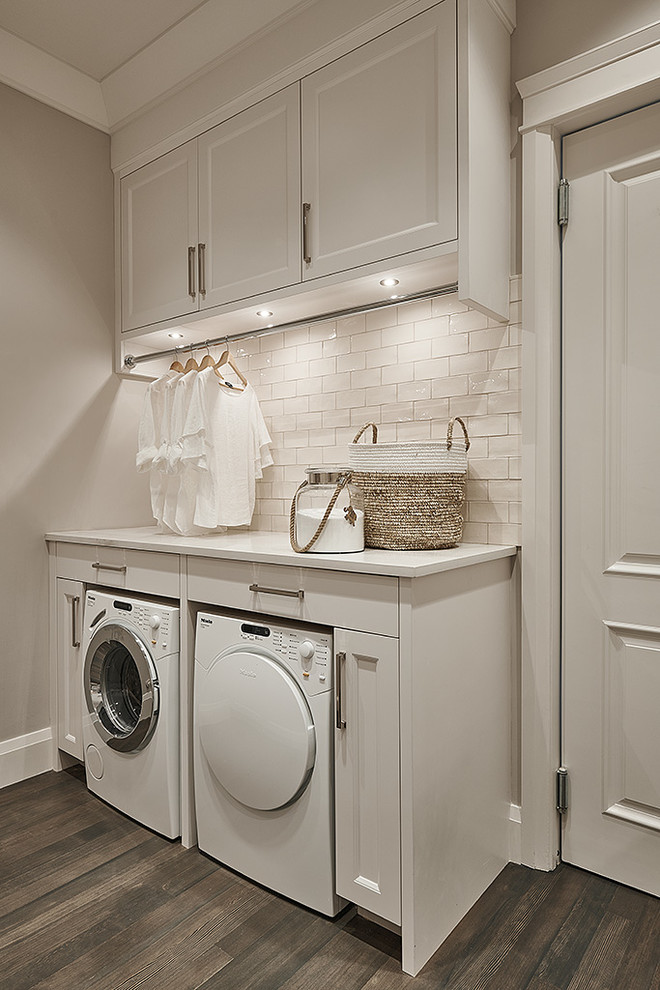 Inredning av en klassisk liten vita vitt tvättstuga enbart för tvätt, med luckor med infälld panel, vita skåp, bänkskiva i kvarts, grå väggar, mörkt trägolv, en tvättmaskin och torktumlare bredvid varandra och brunt golv