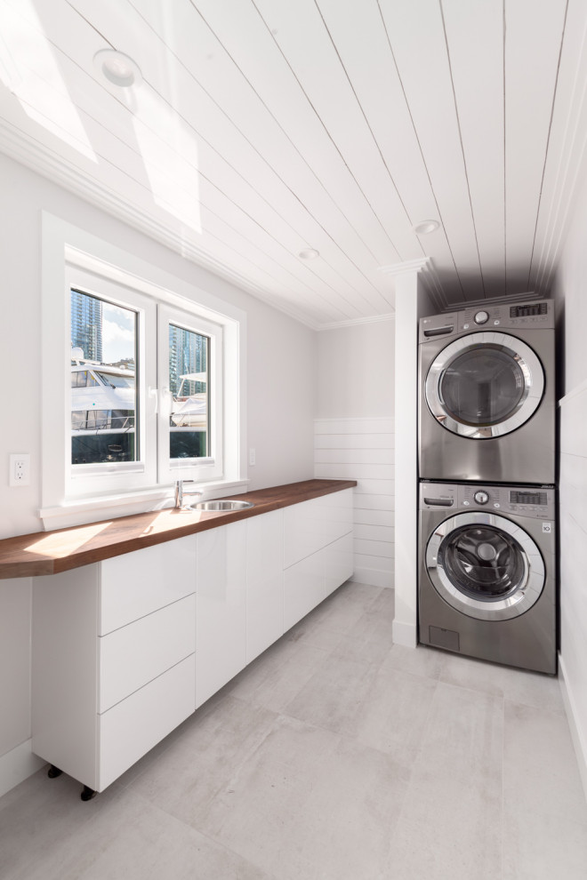 Design ideas for a medium sized classic single-wall utility room in Vancouver with a built-in sink, flat-panel cabinets, white cabinets, wood worktops, white walls, porcelain flooring, a stacked washer and dryer, grey floors, brown worktops and a dado rail.