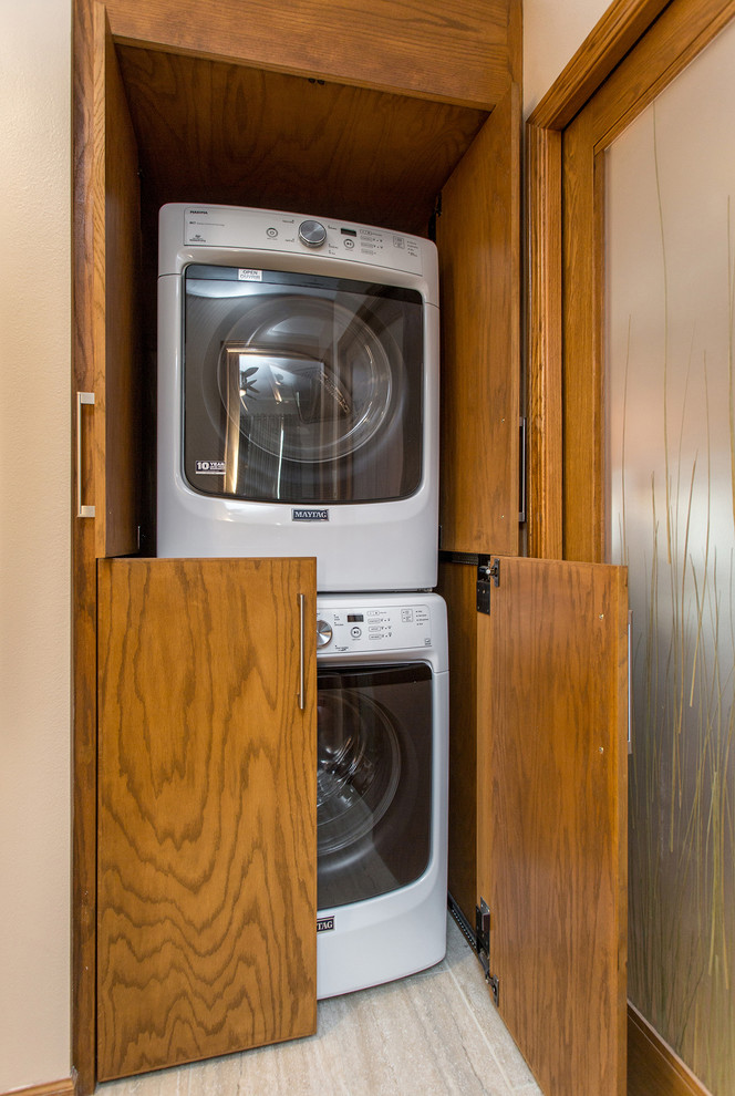 Modelo de armario lavadero clásico renovado pequeño con paredes beige, suelo de madera oscura, armarios con paneles lisos, puertas de armario de madera oscura y lavadora y secadora apiladas