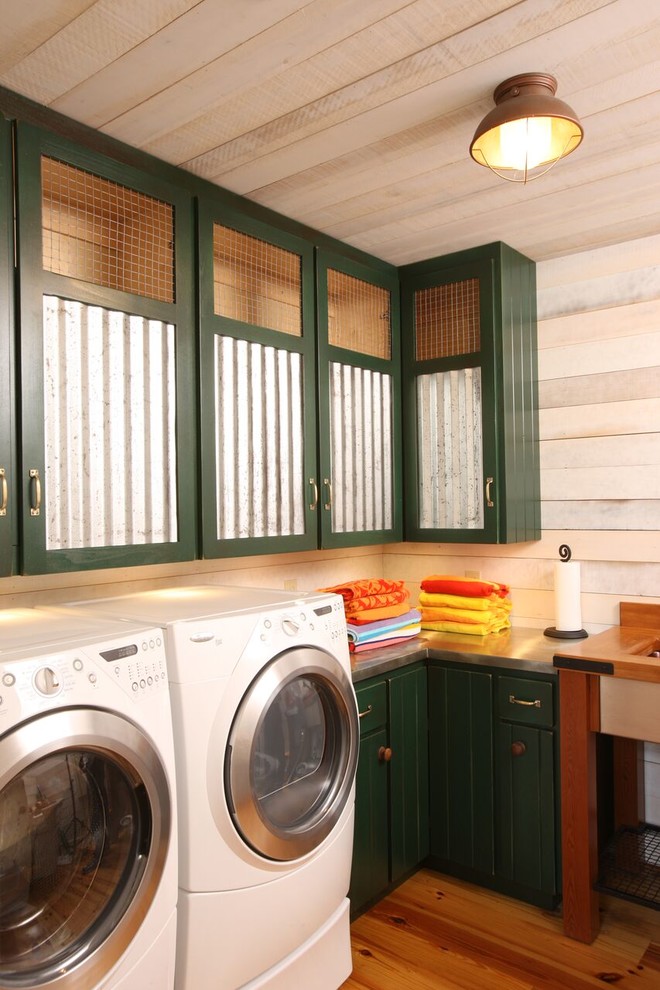 Idéer för en stor rustik l-formad tvättstuga enbart för tvätt, med gröna skåp, vita väggar, en tvättmaskin och torktumlare bredvid varandra, bänkskiva i rostfritt stål och mellanmörkt trägolv