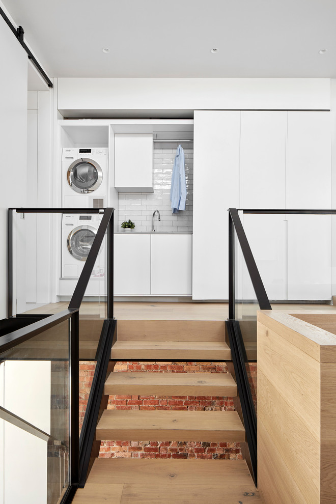 Exempel på en liten modern linjär liten tvättstuga, med släta luckor, vita skåp, bänkskiva i kvarts, vita väggar, ljust trägolv, en tvättpelare och beiget golv