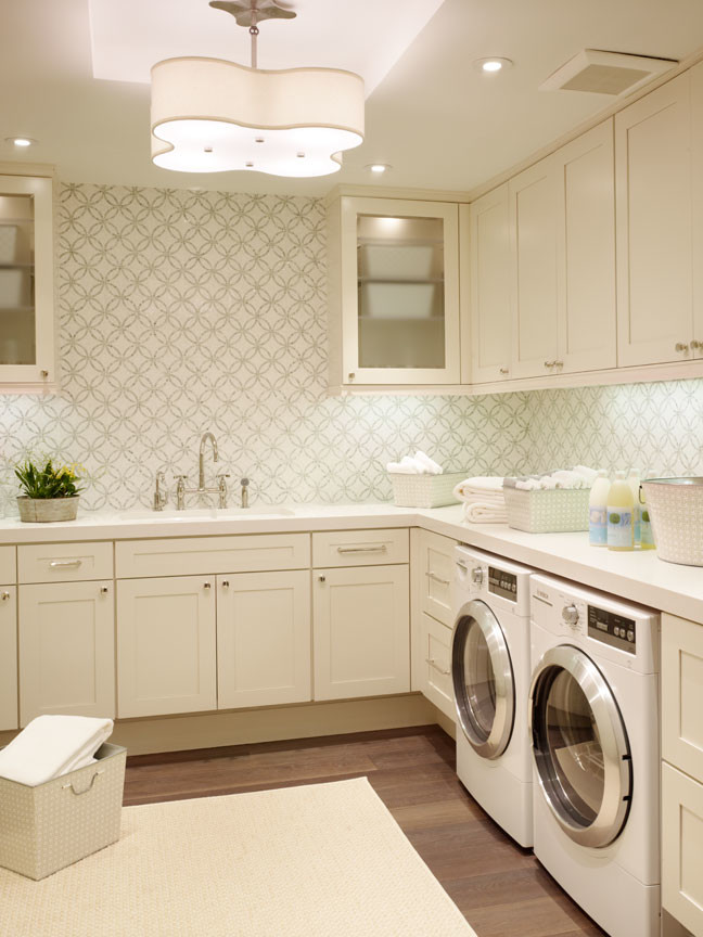 Modern inredning av en l-formad tvättstuga enbart för tvätt, med brunt golv, en integrerad diskho, skåp i shakerstil, vita skåp, vita väggar, mellanmörkt trägolv och en tvättmaskin och torktumlare bredvid varandra
