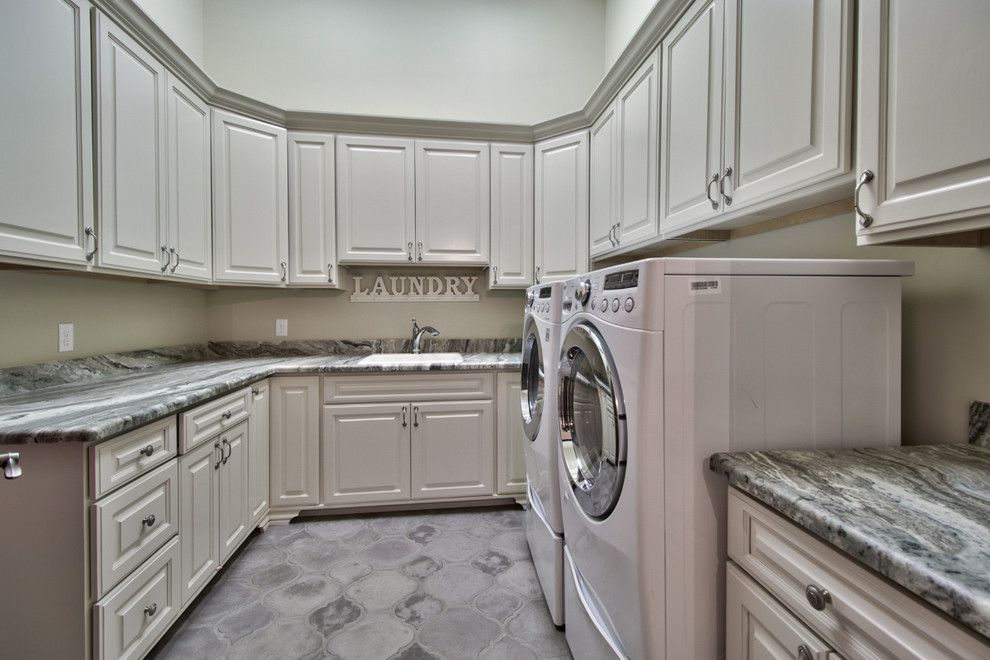 Klassisk inredning av en stor grå u-formad grått tvättstuga enbart för tvätt, med en nedsänkt diskho, luckor med upphöjd panel, vita skåp, granitbänkskiva, grå väggar, betonggolv, en tvättmaskin och torktumlare bredvid varandra och grått golv