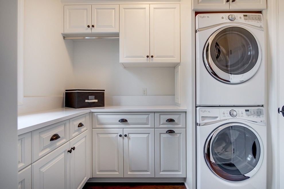 Klassisk inredning av en l-formad tvättstuga enbart för tvätt, med vita skåp, bänkskiva i kvartsit, vita väggar, mörkt trägolv och en tvättpelare