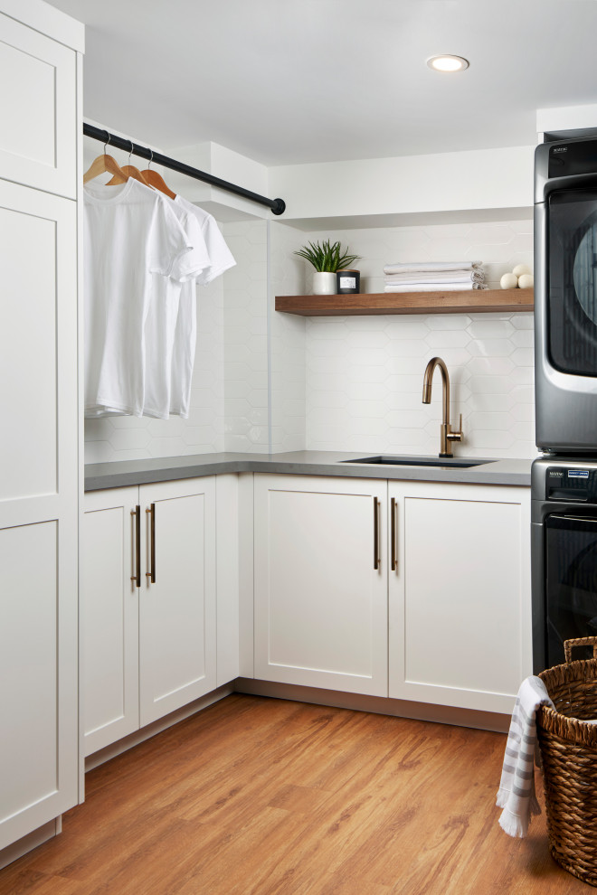 Modern inredning av en mellanstor grå l-formad grått tvättstuga enbart för tvätt, med en enkel diskho, skåp i shakerstil, vita skåp, bänkskiva i kvarts, vitt stänkskydd, stänkskydd i porslinskakel, vita väggar, mellanmörkt trägolv, en tvättpelare och brunt golv