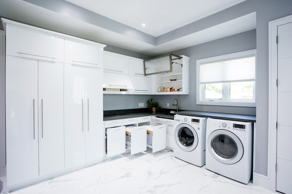 Bild på en stor funkis grå l-formad grått tvättstuga enbart för tvätt, med en undermonterad diskho, släta luckor, vita skåp, bänkskiva i kvarts, grå väggar, marmorgolv, en tvättmaskin och torktumlare bredvid varandra och vitt golv