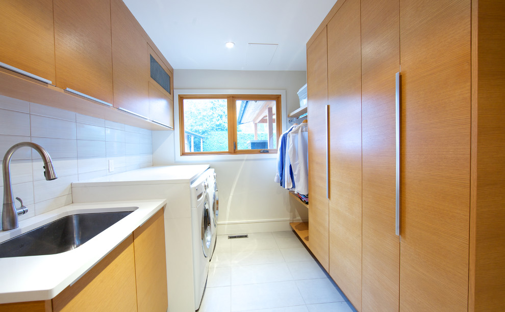 Zweizeilige, Große Moderne Waschküche mit Unterbauwaschbecken, flächenbündigen Schrankfronten, hellbraunen Holzschränken, Waschmaschine und Trockner nebeneinander, weißer Wandfarbe und Keramikboden in Vancouver