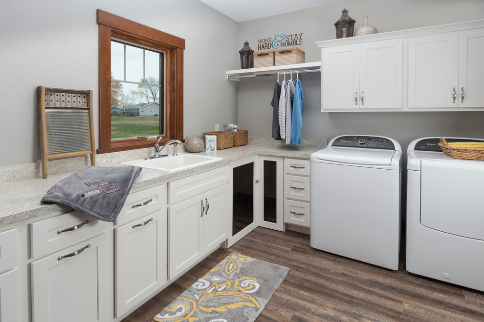 Example of a mid-sized classic l-shaped medium tone wood floor and brown floor dedicated laundry room design in Other with shaker cabinets, white cabinets, a drop-in sink, solid surface countertops, gray walls and a side-by-side washer/dryer